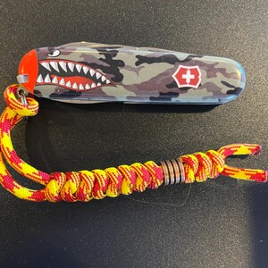 May include: A multi-tool knife with a camouflage pattern and a red and white shark mouth design. The knife has a red and yellow lanyard attached. The Swiss cross logo is visible on the side.