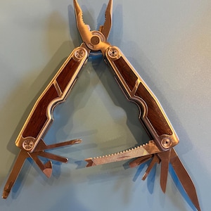 May include: A multi-tool with a stainless steel finish and dark wood grain handles. The tool features pliers, a saw, a screwdriver, and other implements. The tool is open, displaying its various functions.