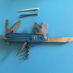 May include: A blue Swiss Army knife with multiple tools, including a blade, scissors, and corkscrew. The knife has a transparent blue handle and a silver cross emblem. A toothpick and tweezers are also visible.
