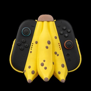 May include: A unique yellow banana-shaped accessory for a gaming console, with brown spots and a wooden top. The accessory is flanked by two black controllers with colored buttons. The background is solid black.