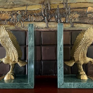 May include: A pair of bookends featuring a gold-colored eagle with outstretched wings, mounted on a green marble base. The eagle is detailed with textured feathers. The background shows a relief of a Western scene.