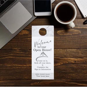 May include: A door hanger with the text "Welcome to our Open House!" in a script font. The door hanger has a marble-like pattern and includes instructions to remove shoes and sign in. A laptop, phone, and coffee cup are in the background.