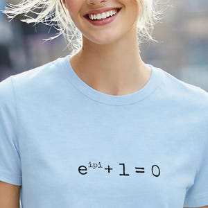 Op de afbeelding: Een lichtblauw T-shirt met korte mouwen en de wiskundige vergelijking e^ipi + 1 = 0 in zwarte letters op het midden van de borst gedrukt. Het kledingstuk heeft een ronde hals en een standaard pasvorm, gemaakt van een effen stof.