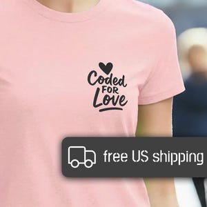 Puede incluir: Camiseta rosa claro con la frase "Coded for Love" y un corazón. La camiseta de manga corta tiene cuello redondo. La persona de la foto sonríe al aire libre.