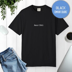 Peut inclure: Un t-shirt noir &agrave; manches courtes avec le texte Amor fati imprim&eacute; en blanc au centre de la poitrine. Le t-shirt a une encolure ronde et une &eacute;tiquette au col int&eacute;rieur indiquant Comfort Colors. Un badge circulaire bleu affiche Black Comfort Colors.