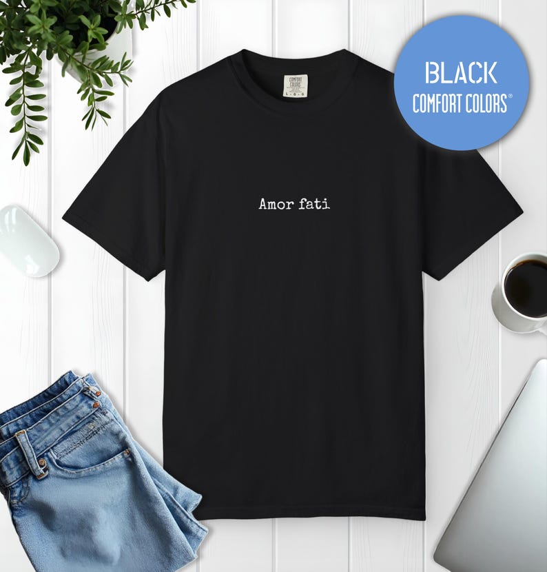 Peut inclure: Un t-shirt noir &agrave; manches courtes avec le texte Amor fati imprim&eacute; en blanc au centre de la poitrine. Le t-shirt a une encolure ronde et une &eacute;tiquette au col int&eacute;rieur indiquant Comfort Colors. Un badge circulaire bleu affiche Black Comfort Colors.