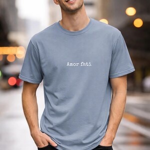 Peut inclure: Un t-shirt &agrave; manches courtes bleu poussi&eacute;reux avec le texte "Amor fati" imprim&eacute; en blanc dans une police de style machine &agrave; &eacute;crire au centre de la poitrine. Le v&ecirc;tement pr&eacute;sente un col rond classique et une coupe standard en tissu uni.