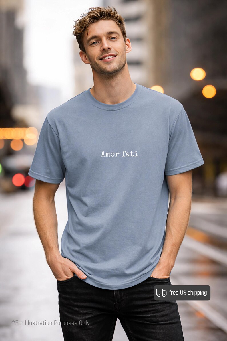 Peut inclure: Un t-shirt &agrave; manches courtes bleu poussi&eacute;reux avec le texte "Amor fati" imprim&eacute; en blanc dans une police de style machine &agrave; &eacute;crire au centre de la poitrine. Le v&ecirc;tement pr&eacute;sente un col rond classique et une coupe standard en tissu uni.