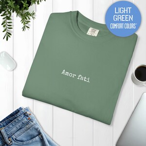 Peut inclure: Un t-shirt &agrave; col rond vert clair avec le texte "Amor fati" brod&eacute; en fil blanc au centre de la poitrine. Le t-shirt pr&eacute;sente un col c&ocirc;tel&eacute; et une &eacute;tiquette de la marque Comfort Colors visible &agrave; l'int&eacute;rieur de l'encolure.