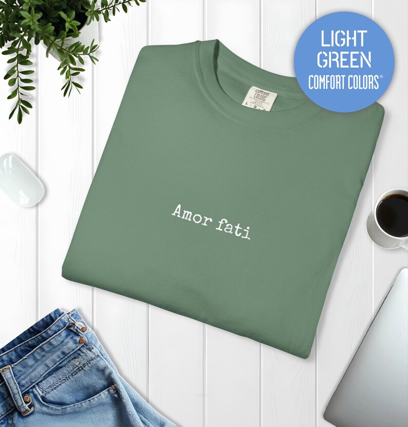 Peut inclure: Un t-shirt &agrave; col rond vert clair avec le texte "Amor fati" brod&eacute; en fil blanc au centre de la poitrine. Le t-shirt pr&eacute;sente un col c&ocirc;tel&eacute; et une &eacute;tiquette de la marque Comfort Colors visible &agrave; l'int&eacute;rieur de l'encolure.