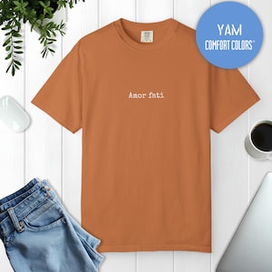 Peut inclure: Un t-shirt &agrave; manches courtes et col rond d'une couleur orange br&ucirc;l&eacute; unie. Le centre de la poitrine pr&eacute;sente le texte "Amor fati" imprim&eacute; dans une police blanche simple. L'&eacute;tiquette int&eacute;rieure du col affiche la marque Comfort Colors.