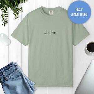 Peut inclure: Un t-shirt &agrave; manches courtes vert sauge avec le texte Amor fati imprim&eacute; en noir dans une police de style machine &agrave; &eacute;crire au centre de la poitrine. Le v&ecirc;tement pr&eacute;sente une encolure ronde et une &eacute;tiquette de marque Comfort Colors au col.
