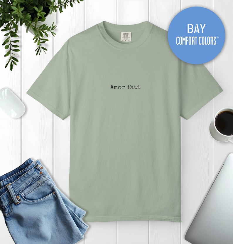 Peut inclure: Un t-shirt &agrave; manches courtes vert sauge avec le texte Amor fati imprim&eacute; en noir dans une police de style machine &agrave; &eacute;crire au centre de la poitrine. Le v&ecirc;tement pr&eacute;sente une encolure ronde et une &eacute;tiquette de marque Comfort Colors au col.