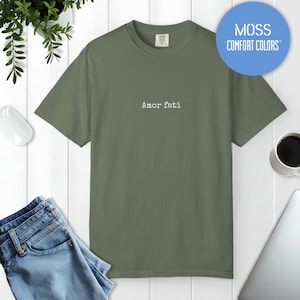 Peut inclure: Un t-shirt &agrave; manches courtes vert mousse avec le texte Amor fati imprim&eacute; en blanc au centre de la poitrine. Le v&ecirc;tement pr&eacute;sente une encolure ronde et une coupe standard. Un graphique circulaire bleu en haut &agrave; droite affiche MOSS COMFORT COLORS.