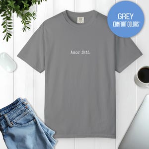 Peut inclure: Un t-shirt gris &agrave; manches courtes avec le texte Amor fati imprim&eacute; en blanc au centre de la poitrine. Le t-shirt pr&eacute;sente une encolure ronde et une coupe standard. Un graphique circulaire bleu en haut &agrave; droite contient le texte GREY COMFORT COLORS.