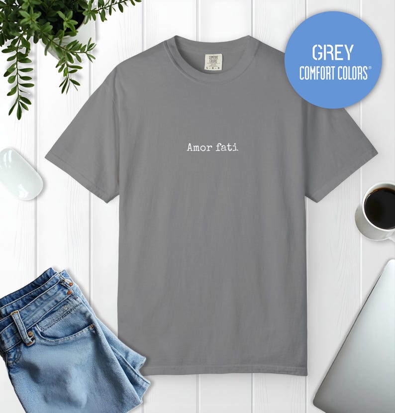 Peut inclure: Un t-shirt gris &agrave; manches courtes avec le texte Amor fati imprim&eacute; en blanc au centre de la poitrine. Le t-shirt pr&eacute;sente une encolure ronde et une coupe standard. Un graphique circulaire bleu en haut &agrave; droite contient le texte GREY COMFORT COLORS.