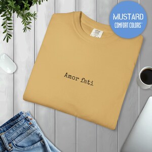 Peut inclure: Un t-shirt &agrave; col rond jaune moutarde avec le texte "Amor fati" brod&eacute; en fil noir au centre de la poitrine. Le t-shirt pr&eacute;sente un col c&ocirc;tel&eacute; et une &eacute;tiquette de la marque Comfort Colors visible &agrave; l'int&eacute;rieur du col.