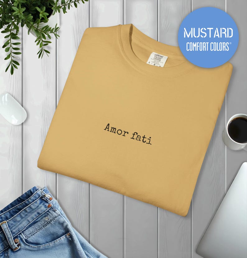Peut inclure: Un t-shirt &agrave; col rond jaune moutarde avec le texte "Amor fati" brod&eacute; en fil noir au centre de la poitrine. Le t-shirt pr&eacute;sente un col c&ocirc;tel&eacute; et une &eacute;tiquette de la marque Comfort Colors visible &agrave; l'int&eacute;rieur du col.