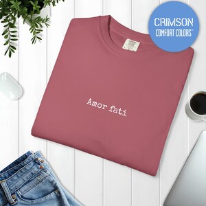Peut inclure: Un t-shirt &agrave; col rond de couleur cramoisi avec le texte "Amor fati" brod&eacute; en fil blanc au centre de la poitrine. Le v&ecirc;tement pr&eacute;sente une &eacute;tiquette de marque Comfort Colors &agrave; l'int&eacute;rieur du col et un logo circulaire bleu.