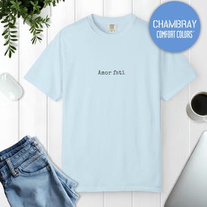 Peut inclure: Un t-shirt bleu clair &agrave; manches courtes avec le texte Amor fati imprim&eacute; en police noire de style machine &agrave; &eacute;crire au centre de la poitrine. Le v&ecirc;tement pr&eacute;sente une encolure ronde et une coupe standard, avec une &eacute;tiquette visible au col.