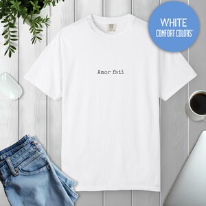 Peut inclure: Un t-shirt blanc &agrave; manches courtes avec un col rond, pr&eacute;sentant le texte Amor fati imprim&eacute; dans une police noire de style machine &agrave; &eacute;crire au centre de la poitrine. Le t-shirt a une coupe standard et est de la marque Comfort Colors.