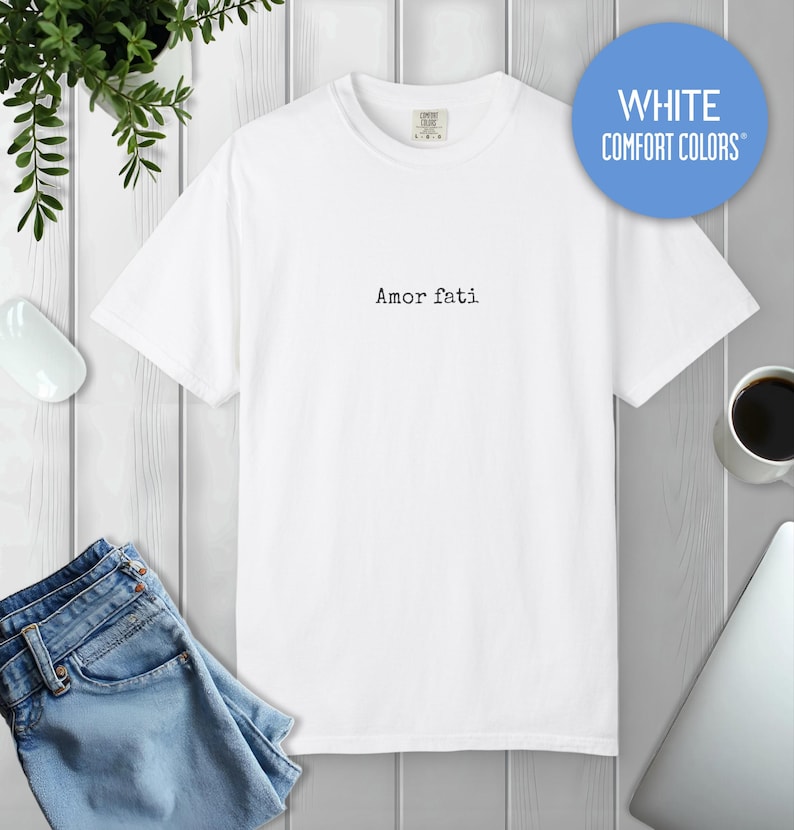 Peut inclure: Un t-shirt blanc &agrave; manches courtes avec un col rond, pr&eacute;sentant le texte Amor fati imprim&eacute; dans une police noire de style machine &agrave; &eacute;crire au centre de la poitrine. Le t-shirt a une coupe standard et est de la marque Comfort Colors.