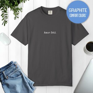 Peut inclure: Un t-shirt gris fonc&eacute; &agrave; manches courtes avec le texte Amor fati imprim&eacute; en police de style machine &agrave; &eacute;crire blanche sur le centre de la poitrine. Le t-shirt pr&eacute;sente un col rond et une &eacute;tiquette au col int&eacute;rieur indiquant Comfort Colors.