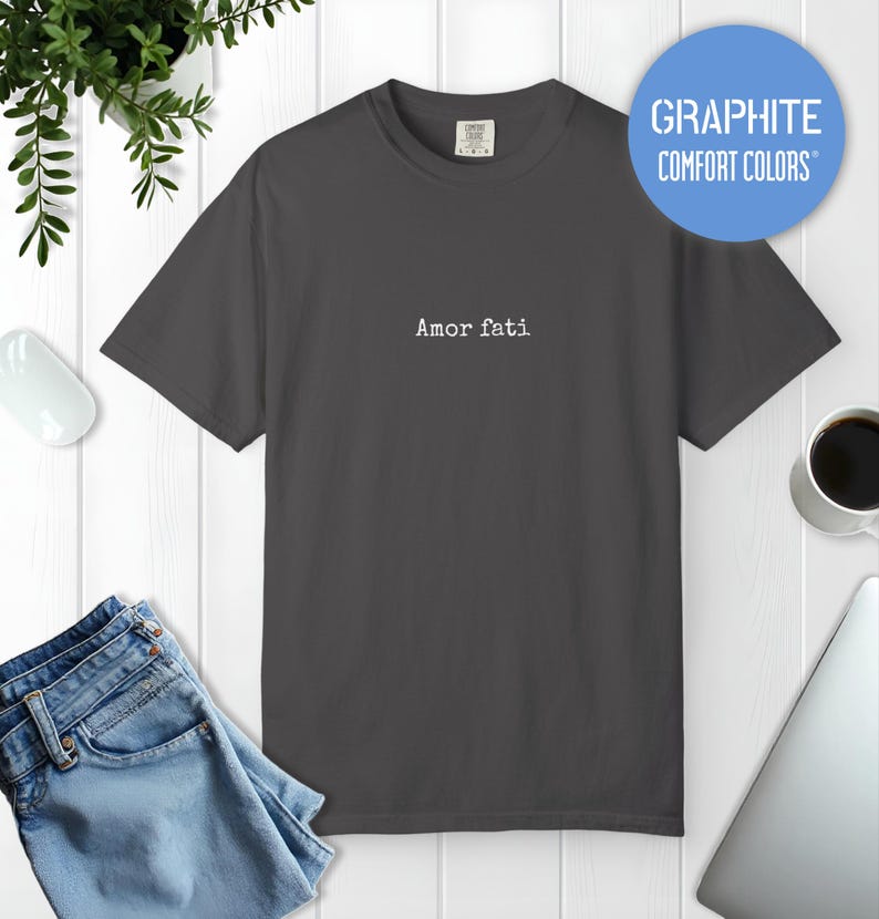 Peut inclure: Un t-shirt gris fonc&eacute; &agrave; manches courtes avec le texte Amor fati imprim&eacute; en police de style machine &agrave; &eacute;crire blanche sur le centre de la poitrine. Le t-shirt pr&eacute;sente un col rond et une &eacute;tiquette au col int&eacute;rieur indiquant Comfort Colors.