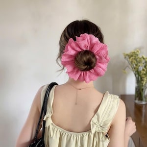 Puede incluir: Un accesorio para el cabello de encaje rosa, que se asemeja a una flor grande, se usa en un moño. El accesorio tiene un diseño detallado y con volantes. La persona lleva un vestido de color claro. Se ve una correa negra de bolso.
