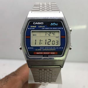 May include: A silver Casio digital watch with a blue and black face. The watch displays the time and date. The watch has a silver strap and is water resistant to 50 metres.