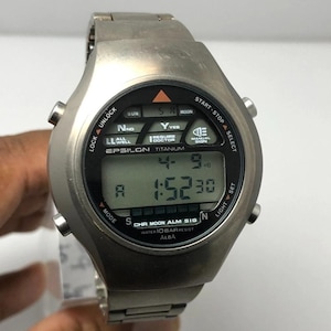 Puede incluir: Un reloj digital plateado con una pantalla negra que muestra la hora y la fecha. La esfera del reloj tiene varios botones y texto, incluyendo "EPSILON TITANIUM" y "WATER 10 BAR RESIST". El reloj tiene una correa de metal plateado.