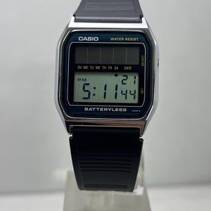 May include: A digital Casio watch with a black strap and silver-tone case. The watch face displays the time, date, and day of the week. The text "CASIO", "WATER RESIST", and "BATTERYLESS" are visible.
