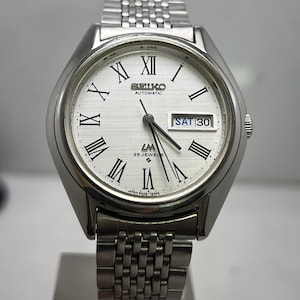 May include: A silver-toned Seiko automatic wristwatch with a white face, black Roman numerals, and a day-date display. The watch features a stainless steel band and a round case. The watch face reads "SEIKO AUTOMATIC" and "LM 25 JEWELS".