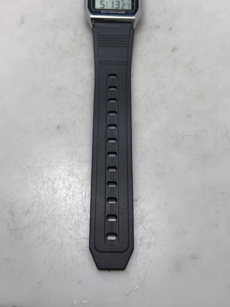 May include: A digital wristwatch with a black resin band. The watch face displays the time, 5:13, and other information. The band has a series of oval holes for adjustment.