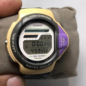 May include: A Casio digital watch with a beige and black case, purple accents, and a digital display showing the time. The watch features an alarm and chronograph functions. The watch is water-resistant.