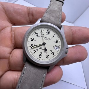 Może przedstawiać: Srebrny zegarek kwarcowy Hamilton Khaki z jasną tarczą i szarym sk&oacute;rzanym paskiem. Tarcza zegarka wyświetla godzinę z oznaczeniami 12- i 24-godzinnymi oraz okienkiem daty. Pasek jest w neutralnym szarym kolorze.
