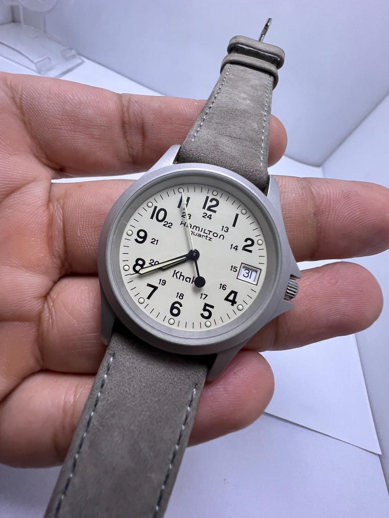 Może przedstawiać: Srebrny zegarek kwarcowy Hamilton Khaki z jasną tarczą i szarym sk&oacute;rzanym paskiem. Tarcza zegarka wyświetla godzinę z oznaczeniami 12- i 24-godzinnymi oraz okienkiem daty. Pasek jest w neutralnym szarym kolorze.