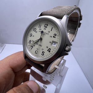 Może przedstawiać: Srebrny zegarek kwarcowy Hamilton Khaki z kremową tarczą i brązowym sk&oacute;rzanym paskiem. Zegarek posiada czarne cyfry, wyświetlacz daty i nazwę marki. Zegarek jest prezentowany na przezroczystym stojaku.