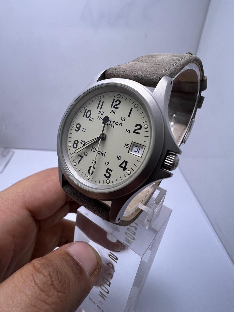 Może przedstawiać: Srebrny zegarek kwarcowy Hamilton Khaki z kremową tarczą i brązowym sk&oacute;rzanym paskiem. Zegarek posiada czarne cyfry, wyświetlacz daty i nazwę marki. Zegarek jest prezentowany na przezroczystym stojaku.