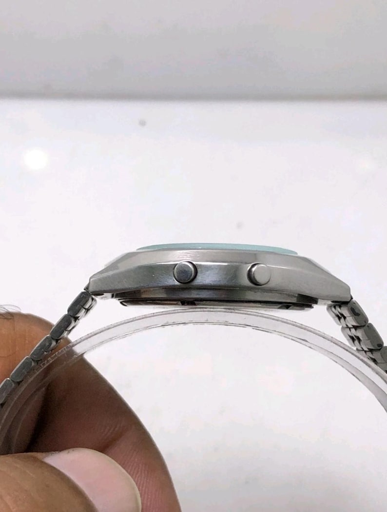 May include: A silver-toned wristwatch with a light blue face and a silver metal band. The watch has two buttons on the side and a clear plastic band holder. The watch is a classic style.