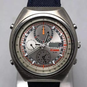 May include: Silver-toned K288 ALBA chronograph watch with a black fabric band. The watch face features multiple dials, red and orange accents, and the number 12 prominently displayed. The watch has a tachymeter scale.