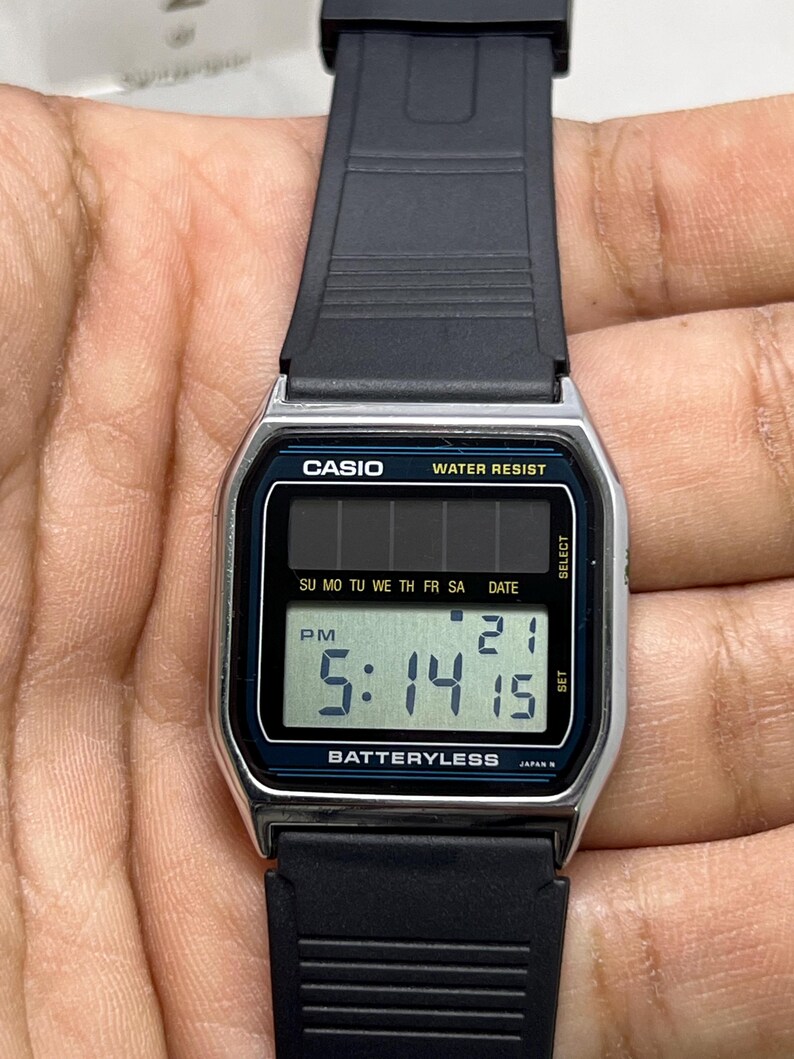 May include: A Casio digital watch with a black strap and a silver-colored face. The watch displays the time as 5:14:15 PM and the date as 21. The watch face has the text "CASIO", "WATER RESIST", and "BATTERYLESS".