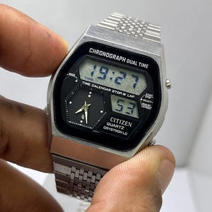 Könnte beinhalten: Eine silberne Citizen Quartz Crystron LC Digitaluhr mit schwarzem Zifferblatt und silbernem Armband. Die Uhr zeigt die Uhrzeit 19:27 und 53 an. Die Uhr trägt den Text "CHRONOGRAPH DUAL TIME" und "TIME CALENDAR STOP.W LAP".