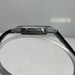 May include: A side view of a digital watch with a black leather band. The watch has a silver-colored case and a dark display. The band is attached to the watch case with small silver pins.