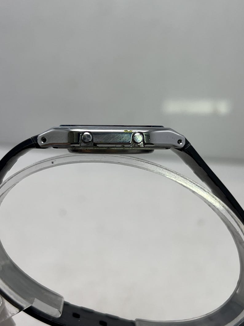 May include: A side view of a digital watch with a black leather band. The watch has a silver-colored case and a dark display. The band is attached to the watch case with small silver pins.