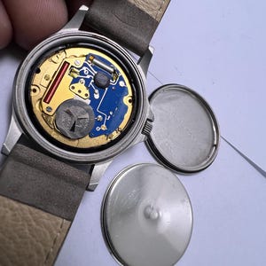 Może przedstawiać: Otwarty zegarek na rękę z szarym sk&oacute;rzanym paskiem, odsłaniający wewnętrzne elementy. Tarcza zegarka jest srebrna z niebieską płytką drukowaną, złotymi akcentami i srebrną baterią. Tylna pokrywa jest odłączona.