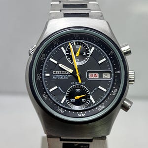 May include: A silver-toned Citizen Chronograph automatic watch with a black face. The watch features yellow hands and markers, a day and date display, and a stainless steel band. The watch face displays the text "CITIZEN CHRONOGRAPH AUTOMATIC 23 JEWELS".