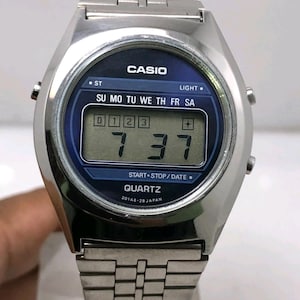 May include: Silver-toned Casio digital watch with a blue face. The watch displays the time as 7:37 and the day of the week. The watch has a stainless steel band and features the text "CASIO" and "QUARTZ".