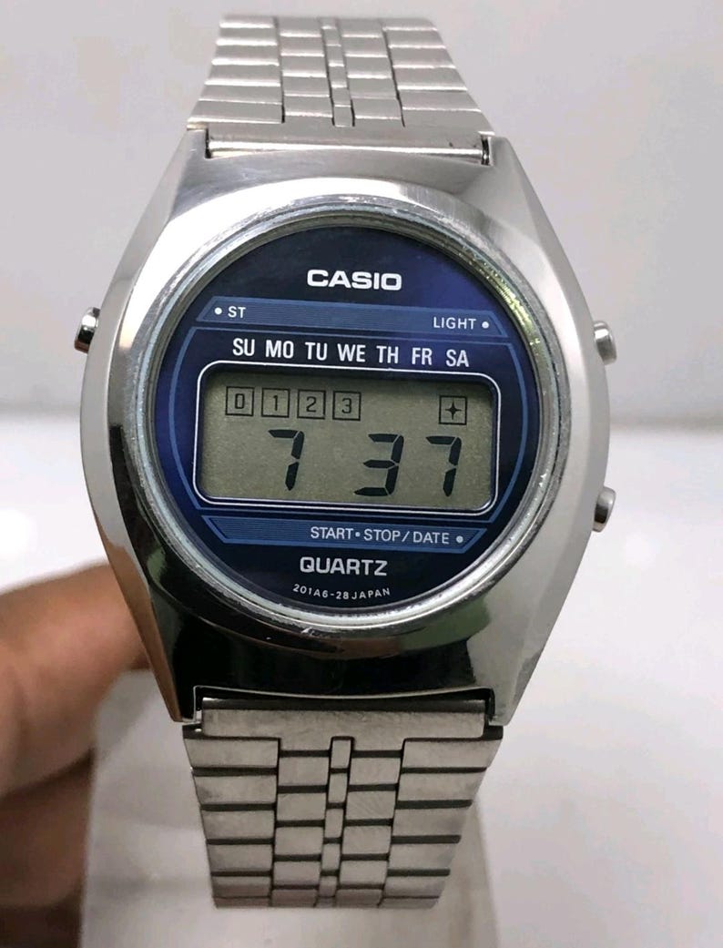 May include: Silver-toned Casio digital watch with a blue face. The watch displays the time as 7:37 and the day of the week. The watch has a stainless steel band and features the text "CASIO" and "QUARTZ".