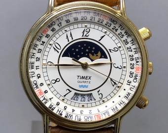 May include: A gold-toned Timex Quartz watch with a white face, featuring a moon phase indicator and day/date display. The watch has a brown leather strap and gold accents. The face displays the days of the week and the date.